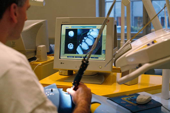 Foto av en mann som sitter med et instrument foran en dataskjerm. På skjermen vises grafiske bilder av tenner.