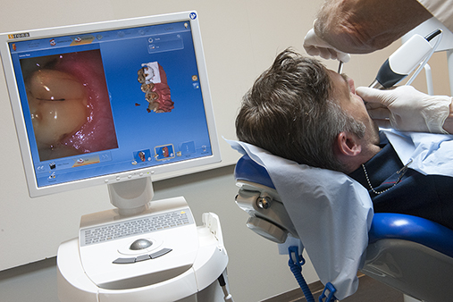 Foto av en pasient som får tennene skannet med håndholdt instrument, og til venstre en dataskjerm som viser bilde av tennene som blir skannet.
