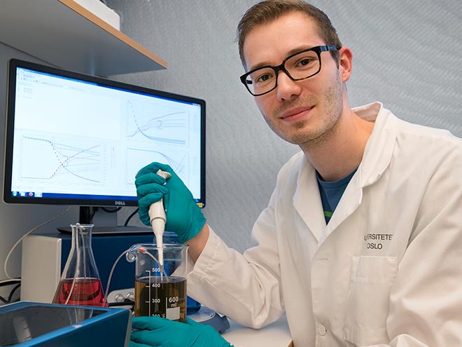 Foto av doktoranden som sitter i en lab med labfrakk foran instrumentene sine.