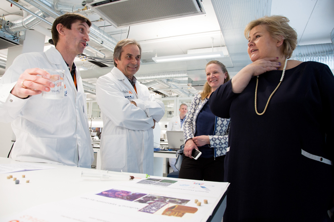 To menn i hvite lab-frakker forklarer noe til statsministeren til høyre, inne på en lab
