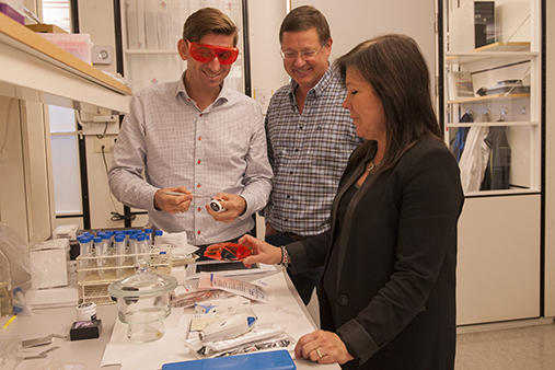 De tre forskerne står i laben og ser på labutstyr, han lengst til venstre har på vernebriller. Alle smiler.
