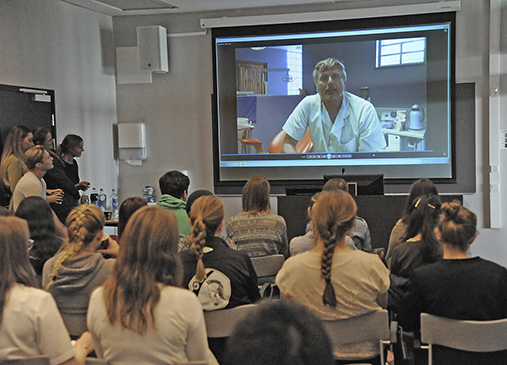 Foto av klasserom med video med doktor som snakker på veggen, mange studenter sitter i klasserommet og ser på.